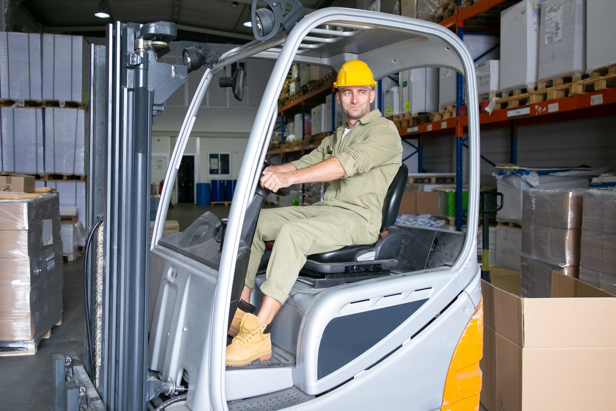 trabajador-logistico-masculino-positivo-en-casco-de-conduccion-de-montacargas-en-almacen-sonriendo-mirando-otro-lado